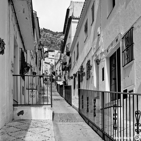 A narrow, picturesque street in Mijas with traditional black wrought iron balconies. The scene features cobblestone pathways and greenery, framed by surrounding hills, creating a serene and charming atmosphere in black and white.