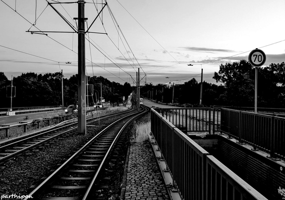 Black and white photo of train tracks and a road running parallel, with a 70 km/h speed limit sign on the right, power lines overhead, and trees in the background.

Schwarzweissfoto von Bahngleisen und einer parallel verlaufenden Strasse, mit einem 70 km/h-Geschwindigkeitsbegrenzungsschild rechts, Stromleitungen ueber ihnen und Baeumen im Hintergrund.