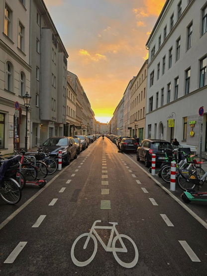Lange, leere Straße mit Fahrradboden-Markierung. Die Sonne geht in der Häuserflucht unter.