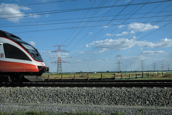 Das Foto zeigt einen modernen Zug, der von links ins Bild fährt. Im Hintergrund ist eine Kulturlandschaft mit Feldern zu sehen. Über dem Zug und der Landschaft verlaufen zahlreiche Stromleitungen, die an hohen Masten befestigt sind. Der Himmel ist blau und weist einige weiße Wolken auf. Die Szene wirkt dynamisch und technisch fortschrittlich.
 --- 
The photo shows a modern train entering the frame from the left. In the background is a cultivated landscape with fields. Numerous power lines, attached to tall poles, run above the train and the landscape. The sky is blue and has a few white clouds. The scene appears dynamic and technologically advanced.