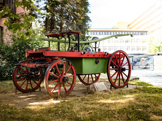 Historische Feuerwehrspritze mit roten Holzrädern und grünem Wassertank, auf einer Wiese zwischen Bäumen und Gebäuden.