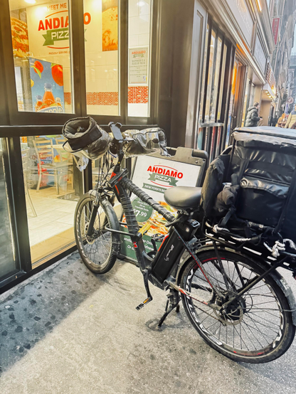 A color photo of an electric bicycle parked outside of a pizza shop on the sidewalk. The bicycle has a frame that is wrapped in black electrical tape and big gloves attached to the handlebars with plastic over them that protect your hands from the wind and cold. There is a mounted phone holder in between the handlebars and a very large insulated carrier bag on the back. It is sitting in front of a sidewalk sign that says Andiamo Pizza. It is up against the windows of the shop and again you can see the words on the Andiamo Pizza on a menu board inside.