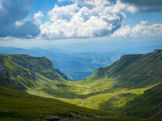 A wide, scenic landscape view looking down a lush green mountain valley under a bright blue sky filled with fluffy white clouds. Sunlight highlights the steep, grassy slopes on either side of the valley floor, where a winding trail or stream runs through the center. Distant blue mountain ranges fade softly into the horizon under shafts of sunlight.