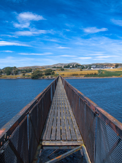 Photo looking down a long, narrow metal footbridge with missing and weathered wooden planks. The bridge stretches straight across a blue lake under a bright blue sky with wispy clouds. Across the bridge yellow and green fields and a small village sit with distant mountains in the background.