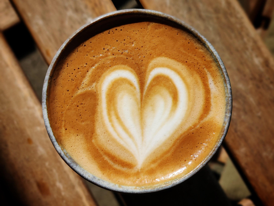A close-up photo of a cafe latte in a ceramic cup, placed on a wooden surface. The coffee has white foam heart on its surface.