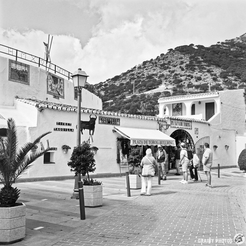 A black and white view of a charming plaza with a bullring, surrounded by palm trees. People gather outside a traditional building, enjoying the architecture and scenery. The background features hills and clouds, adding to the picturesque atmosphere.