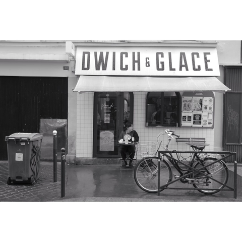 Black and white street photography taken in Paris showing a woman eating a sandwich in a small street with a bike on her side.