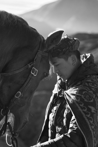 Black and white close up portrait of a horse touching forheads with a man against a mountainous backdrop