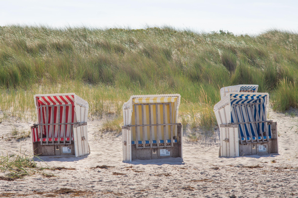 Farbfoto im Querformat. Vier Strandkörbe im Sand, dahinter Dünen. Der linke Korb ist rot-weiß-gestreift, der mittlere gelb-weiß-gestreift und der rechte blau-weiß-gestreift, dahinter lugt noch die Ecke eines weiteren blauen Korbes hervor.