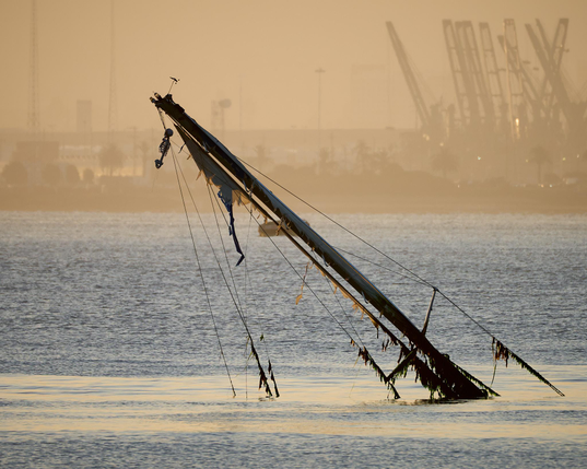 A seaweed laden sailboat mast sticks up out of the water at sunrise. In the distance is the Port of Oakland with cranes reaching high. Hanging from the top of the mast is the head and torso of a skeleton, presumably fake.