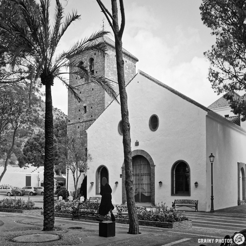 A monochrome image of Iglesia Inmaculada Concepción with a bell tower, surrounded by palm trees and benches in a plaza. A black silhouette figure stands in front of the church, adding a sense of scale to the tranquil scene.
