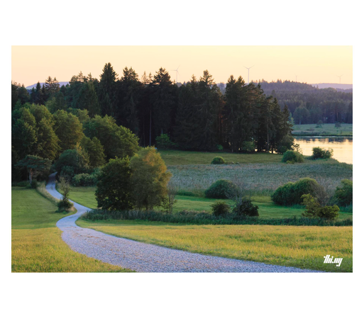 A gravel path leading down a gentle hillside through some fields into the forest surrounding a lake. Yellow golden light & sky. Some wind turbines in the background.