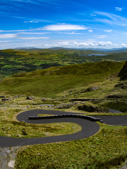 High-angle view of the winding Stwlan Dam access road in Wales. The paved black road twists in a sharp S-curve switchback down a steep, grassy vibrant green mountain. In the distance, rolling green hills, a small lake, and mountain ridges stretch across the horizon under a cloudy blue sky.