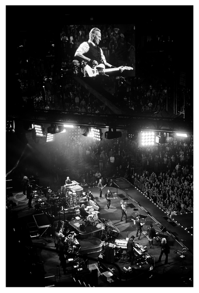 Black and white photo of a Bruce Springsteen concert at the Barclays Center in Brooklyn, taken from the nosebleed seats at the side of the stage. The musicians of the E Street Band appear as tiny figures under the spotlights, facing right and playing to a packed crowd. Above is a giant screen showing a closeup of Bruce in a white shirt and dark vest, strumming his guitar.