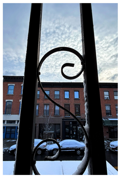 A swirling S-shaped metal strip in between two vertical metal bars, through which we can see a city street in winter. A bit of snow clings to the sides of the bars. Parked cars dusted with snow sit in front of a low-rise brick apartment building with some ground-level storefronts. The wintry blue-white sky is stippled with clouds.