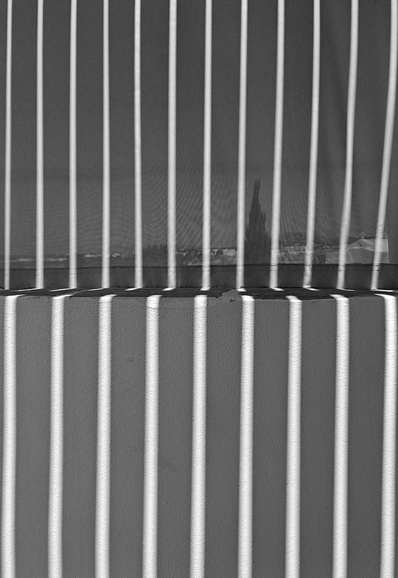 Black and white photo showing  light rays casting vertical stripes onto a semi transparant screen. Below the screen, the same pattern is projected onto a white wall. 