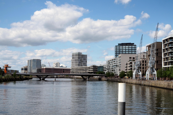 Blick auf moderne Bauten und Hochhäuser am Baakenhafen in der Norderelbe. Im Hintergrund versteckt sich das geschwungene Dach der Elbphilharmonie. Links ein kleiner roter Turm am Ende einer Brücke, rechts alte Kräne, die früher zum Be-und Entladen von Schiffen gedient haben. Der Himmel ist blau, weiße Wolken, das Wasser leicht bewegt.

A view of modern buildings and skyscrapers at Baakenhafen on the northern Elbe. Hidden in the background is the curved roof of the Elbphilharmonie. On the left, a small red tower at the end of a bridge; on the right, old cranes that were once used to load and unload ships. The sky is blue with white clouds, and the water is gently rippling.