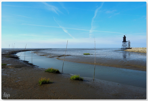 Das Leuchtturmdenkmal bei Obereversand. Im Vordergrund Wasser in einem Priel, abgesteckt mit Birkenzweigen, so genannten Pricken. Der Himmel ist blau, leichte Schleierwolken, das Watt ist braungrau.

The lighthouse monument near Obereversand. In the foreground, water in a tidal channel, marked out with birch twigs, known as ‘Pricken’. The sky is blue with a few wispy clouds; the mudflats are brownish-grey.