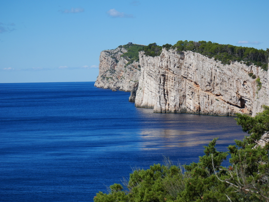 Helle Steilküsten ragen über einem tiefblauen, ruhigen Meer auf. Die Felsen sind oben dicht mit niedrigen Bäumen bewachsen und spiegeln sich stellenweise im glatten Meer, stellenweise aber auch nicht, sodass es aussieht als schwebten manche Abschnitte der Küste, und ein unwirklicher Eindruck entsteht.