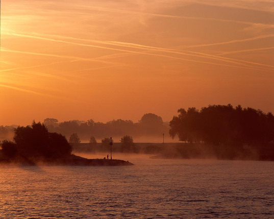 Breiter Fluss mit ruhiger Wasseroberfläche im Vordergrund, im sanften Licht eines orangefarbenen Sonnenaufgangs oder Sonnenuntergangs. Flache Ufer mit vereinzelten Bäumen und niedriger Vegetation, im Hintergrund leichte Nebelschwaden. Am Horizont verlaufen mehrere Kondensstreifen parallel zum Himmel. Die Szene vermittelt die typische Landschaftsstimmung des Niederrheins.