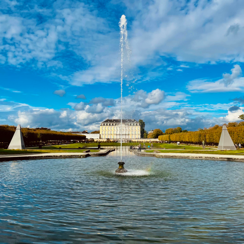 das bild zeigt ein großes barockschloss im hintergrund eines weitläufigen gartens. im vordergrund befindet sich ein rundes wasserbecken mit einer hohen springbrunnenfontäne. der garten ist symmetrisch angelegt mit grünen rasenflächen, getrimmten hecken und zwei pyramidenförmigen objekten an den seiten. über der szenerie erstreckt sich ein intensiv blauer himmel mit vielen weißen wolken.