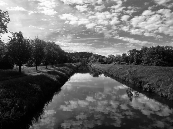 Pfinztal Canal between Grötzingen and Hagsfeld.
A serene black-and-white landscape featuring a straight, tranquil canal running through a countryside. The water creates a clear mirror reflection of a sky filled with scattered, soft cumulus clouds. On the left side, a line of trees casts long shadows across a grassy path. On the right, a sloped bank rises to a line of lush foliage, with low, forested hills visible in the far background.
