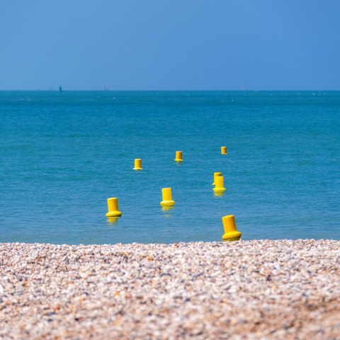 Kieselsteine von einem Strand, dann Meer, dann Himmel. In der Mitte schwimmen sieben gelbe Bojen.