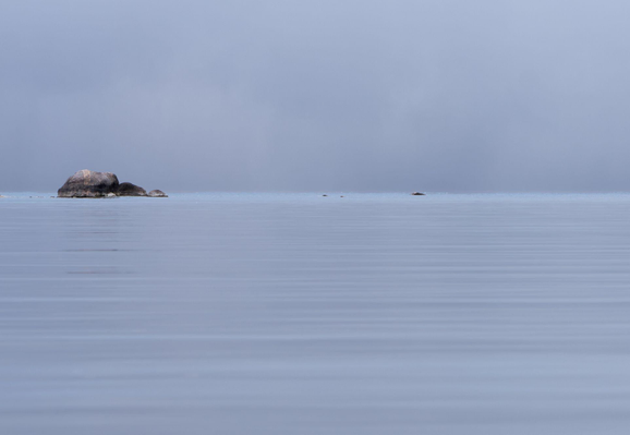 A minimalist image of a light violet-blue still lake with only the slightest ripples and a matching cloudless sky. On the centre left of the frame in the distance, a cluster of rocks sit on the line where the lake meets the sky.