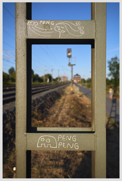 Vertical closeup of a signal tower next to a railway track. The greenish metal beams have been embellished with two small white figures by local street artist peng. Through the opening in the structure the train tracks are visible on the left. On the right, a paved walkway runs parallel to the tracks. A person can be seen on the walkway. The scene is lit by warm late afternoon light.