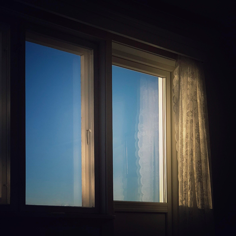 Looking out through two windows at a blue sky from inside. On the right-hand side hangs a white lace curtain, glistening in the sun.