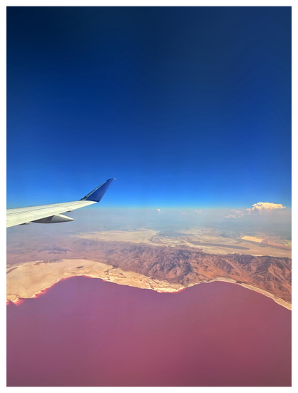 View of Utah’s Great Salt Lake from an airplane window, with the plane’s wing pushing in from left frame. A brown, mountainous landscape sits between the wavy shoreline of a vivid red lake below and a clear blue sky above, with a distant cumulus cloud floating on the horizon. The lake’s red color is caused by the pigmentation of halophilic (salt-loving) bacteria, which thrive in the high salinity of the water.