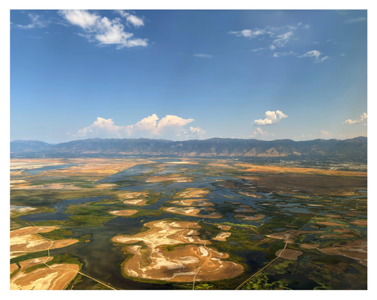 Another view of a Utah landscape from an airplane window. Flat islands of green and brown, connected by a web of narrow causeways, sit in what looks like waterways or a collection of small rivers and lakes — or perhaps these are the drying-up remnants of a larger body of water. Mountains sit at the horizon under a blue sky with clouds.