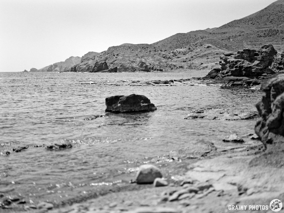 A serene black and white coastal landscape featuring rocky formations and gentle waves lapping at the shore, with distant hills rising in the background under a clear sky.