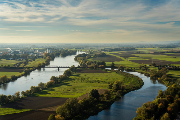 Weite Flusslandschaft aus erhöhter Perspektive. Der Fluss zieht sich in großen Schleifen zwischen Feldern und Baumreihen hindurch; eine schmale Brücke quert ihn. Am Horizont liegt die Landschaft unter einem hellen, wolkigen Himmel.