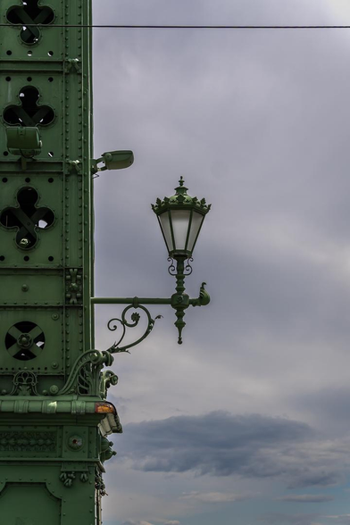 Detailaufnahme einer grün lackierten historischen Eisenkonstruktion. Links füllt ein reich vernietetes Bauteil mit ornamentalen Durchbrüchen das Bild; daraus ragt eine verzierte alte Laterne in den freien Raum. Dahinter ein weiter, grau bewölkter Himmel mit einem schmalen blauen Streifen am Horizont. Eine dünne schwarze Leitung durchquert den oberen Bildrand.