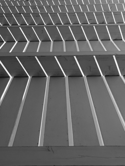 An upside-down black and white photo of a wooden roof with slightly separate planks. Sunlight from above is creating a geometric pattern of parallel lines of light and shadows. 