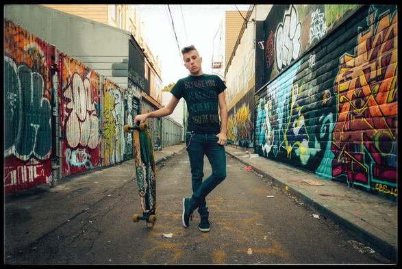 A 20-something skateboarder, in jeans and black T-shirt, stands casually in the center of a graffiti-tagged alley. Legs crossed at the ankles, he is leaning on his long board, with a mostly expressionless, slightly defiant look on his face.