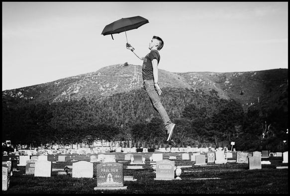 A black and white photo of a young man holding an umbrella, floating through the air above a cemetery.