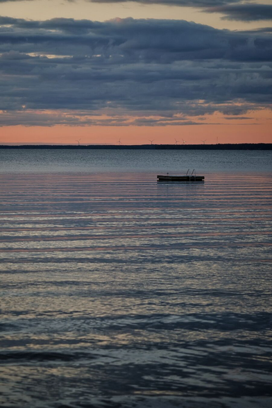 Ein See kurz nach dem Sonnenuntergang, der Himmel ist noch etwas rot aber auch dunkel bewölkt. Man sieht nur das Wasser des Sees, das leichte Wellen schlägt, und das andere Ufer als weit entfernter Strich. Weit auf dem See liegt eine Badeinsel aus Holz mit einer kleinen Leiter. Wenn man genau hinschaut erkennt man so gerade noch eine Möwe, die auf der Badeinsel sitzt.