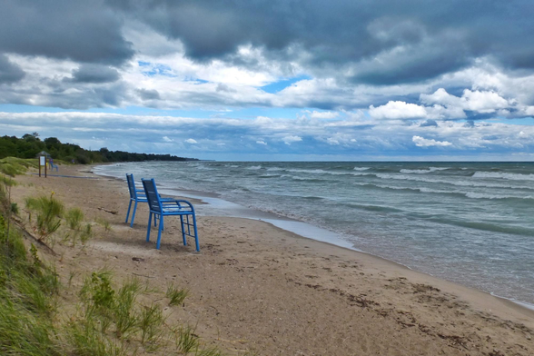 Two very large blue chairs sit on a beach overlooking the rough waters of a large lake. Large waves are seen crashing into the sandy beach and dark moody clouds fill the sky. Trees are seen in the background to the left as well as other small vegetation.
