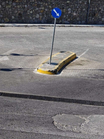A photo showing a small traffic island with a blue traffic sign with a white arrow pointing downward to the right. A yellow painted concrete barrier separates the island from the roads that show patches of repaired asphalt. All on a bright sunny day.