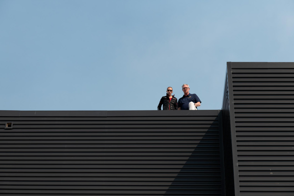 Two people looking ominously down from the roof of a black, horizontally-striped building, set against a blue sky.
