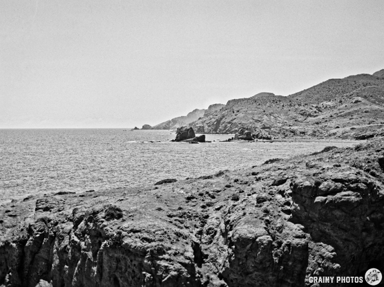 A black and white landscape featuring a rocky shoreline with cliffs and scattered boulders, overlooking a calm sea and distant hills under a clear sky.