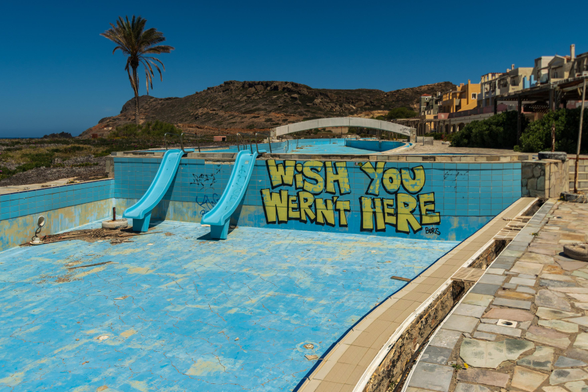 Das Foto zeigt ein verlassenes Schwimmbad, das mit Graffiti übersät ist. Die leeren Becken und die verblassten Farben vermitteln ein Gefühl der Verlassenheit und des Verfalls. Die Graffiti, insbesondere die Botschaft "WISH YOU WERNT HERE", verstärken die melancholische und desillusionierte Stimmung. Die Palmen und die umliegende Landschaft deuten auf ein warmes Klima hin, was den Kontrast zwischen der einst lebendigen Anlage und ihrem jetzigen Zustand noch verstärkt. Es ist ein Bild, das zum Nachdenken über Vergänglichkeit, verpasste Gelegenheiten und die Spuren anregt, die wir hinterlassen.