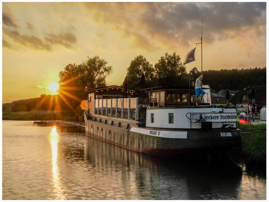 A boat named "Roland" is docked by a river at sunset, with sun rays glimmering on the water. The boat features a restaurant area, and a figure is seen standing on the deck. Surrounding greenery and buildings are visible in the background. 