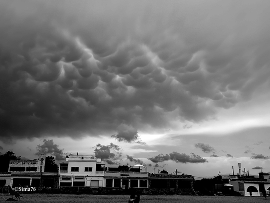 - Photo en noir et blanc d'un ciel d'orage avec des nuages mammatus au-dessus d'un ensemble de bâtiments en bord de mer. 
- Black and white photo of a stormy sky with mammatus clouds above a row of seaside buildings.
- Foto en blanco y negro de un cielo tormentoso con nubes mammatus sobre un conjunto de edificios junto al mar.