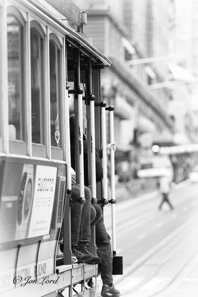 This is a black and white street photo in portrait format of the front of a cable car, packed with tourists in downtown San Francisco, California (2015).

Extending from the right edge to the centre of the image is our cable car (street car or tram). Only the wooden upper part of the cable car is visible, the wheels and chassis are out of view below the photo. It is viewed close up and with the right side in view. The cable car is filling the entire height of the photo, half the width and viewed from a metre or two away. The front portion is open with the legs and feet of the seated passengers visible hanging over the side. Between the seated passengers are vertical wooden posts supporting the roof with white poles attached to the outside enabling any passengers standing on the foot platforms stretching along the sides: to 'hang on for dear life'. Behind the open section is the enclosed cabin, made of wood at the base and with vertical rectangular windows above. On the wooden side are a few simple adverts, blurred slightly out of focus. Ahead of the cable car, the parallel set of rail tracks stretch ahead with the central metal groove hiding the all important moving cable. To the left is a further set of tracks. This and the background are heavily blurred out of focus and slightly overexposed to white. Just visible is a man, perhaps five metres away, crossing the street. Again, ahead of the cable car, on the left side of the road is a line of buildings, the uppers floors out of view above the top of the photo. 