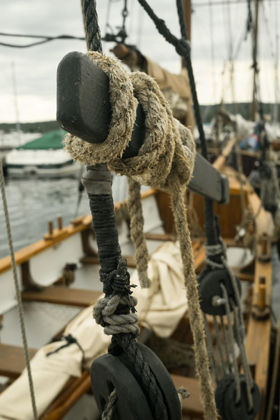 A detail of a nineteenth-century boat in a harbour, focusing on a prickly beige rope twisted around a black painted piece of wood, part of an elaborate black and beige roped rigging. Below the rope is the body of the boat with light, warm wood trim, a white interior, wooden seats and a rolled-up sail. In the background, the water of the harbour, blurred boats, and a green, treed line of land in the distance.