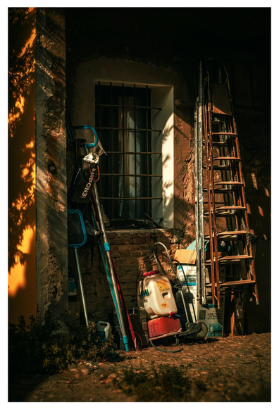 This photo is of a friend's outdoor storage area (it's under an arch) against the exterior wall of a building. On the left-hand side, there are various long-handled pool cleaning nets and tools which lean against yellow wall dappled with leaf shadows. Behind them to the right-ish is a barred window. Underneath the window there is a a few things a red and white portable sprayer tank and boxes etc. on the right there are two tall ladders. One is metal and one is wooden, they are leaning on the weathered stone building. The photograph is filled with warm evening sunglight and a collection of items of a life filled with love.
