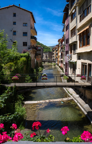 Color photo of a shallow stream running through a Catalan town in the Pyrenees with buildings close on either side and two small bridges spanning the stream.